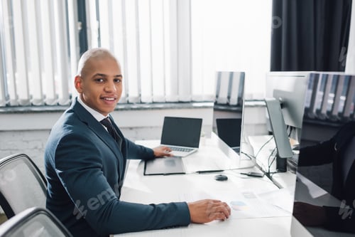Preview: handsome young african american businessman sitting at workplace and smiling at camera in office