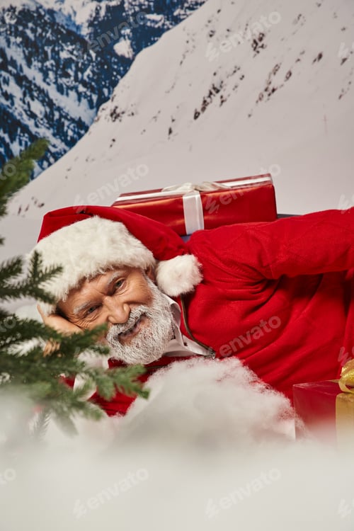 Preview: close up of joyous Santa in christmassy hat lying on snow surrounded by presents, winter concept