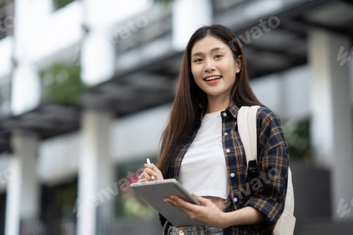 Vista previa: Jovencita asiática segura de sí misma usando una tableta para su proyecto universitario mientras está de pie frente al cl