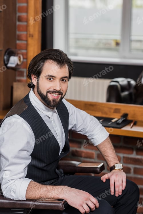 Preview: handsome barber sitting on chair at workplace and looking at camera