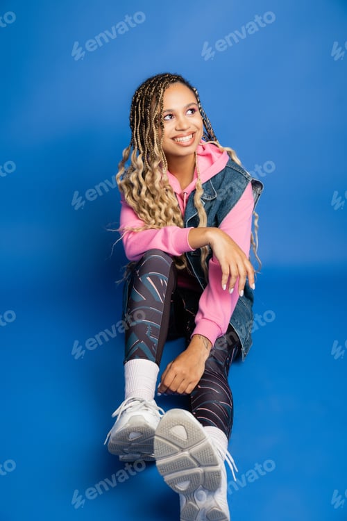 Preview: happy african american woman with dreadlocks sitting on blue background and looking away, sporty