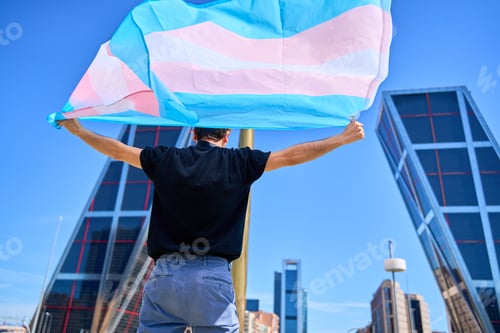 Preview: A man holding a transgender flag in front of a building. The flag is large and colorful.