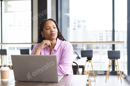 Preview: Woman sitting at table in modern coworking space, gazing thoughtfully with laptop and coffee cup