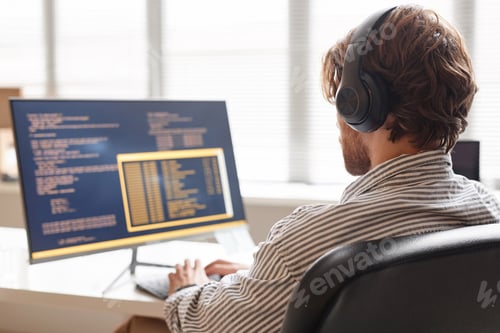 Preview: Male IT Developer Using Computer at Desk in Office and Wearing Headphones