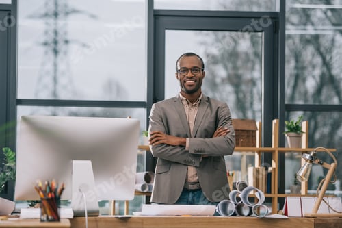 Preview: smiling african american architect with crossed arms standing at workplace with computer and