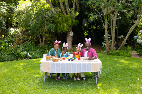 Preview: Happy african american siblings and grandparents in bunny ears painting easter eggs in backyard