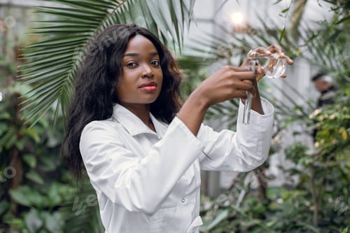 Preview: african woman biologist in white coat pouring clear liquid from glass flask into test tube