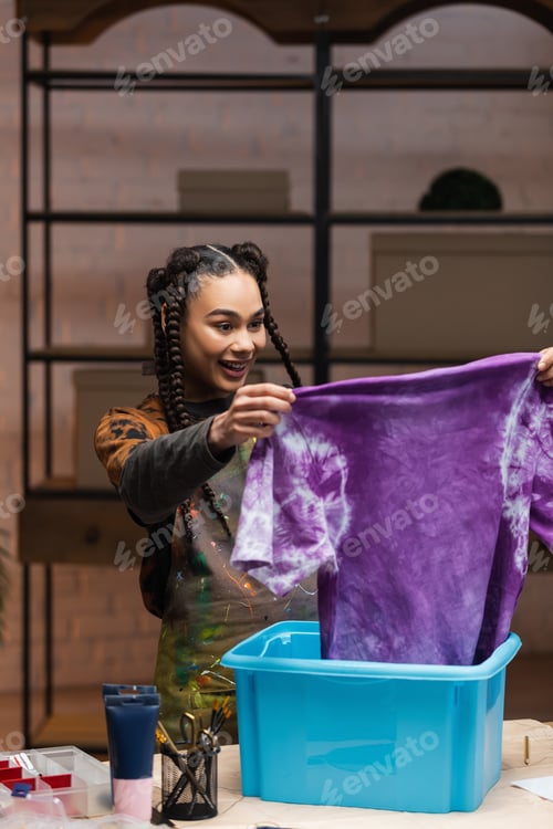 Preview: Positive african american craftswoman holding t-shirt in tie dye style near box in workshop
