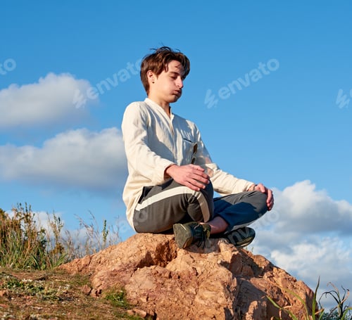 Preview: Shallow focus of a Spanish young male sitting on a rock and meditating against the cloudy blue sky