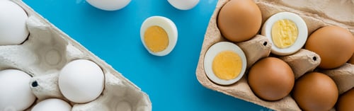 Preview: Top view of boiled and raw eggs near carton containers on blue surface, banner