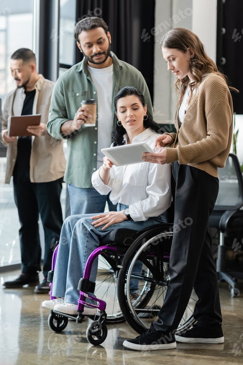 Preview: disabled woman in wheelchair looking at tablet, discussing startup project with interracial team
