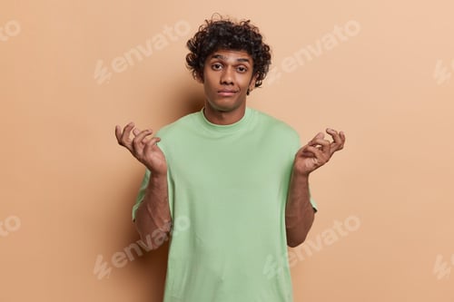 Preview: Waist up shot of clueless curly haired Hindu man spreads palms shrugs shoulders feels unaware wears