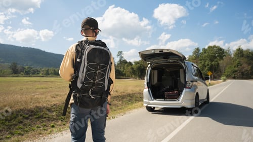 Preview: Smiling young man with backpack opening a trunk door of his car at countryside with blue sky