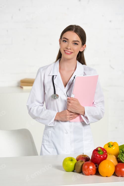 Preview: smiling attractive dietitian in white coat with equipment holding folder near table with fresh