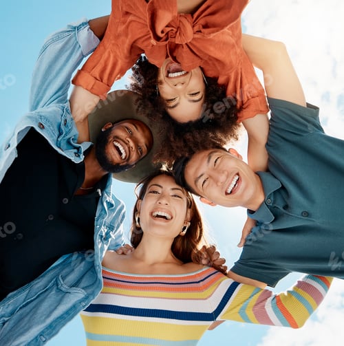 Preview: Cheerful group of diverse friends standing in a huddle against a blue sky. Multi-racial millennial