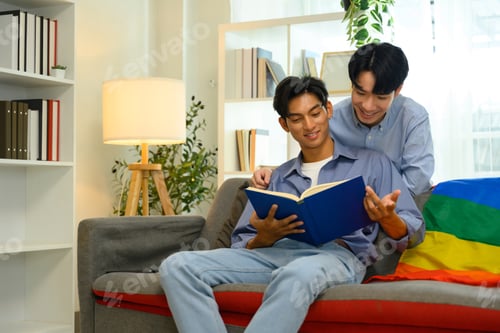 Preview: Happy male gay couple enjoying quality time together and reading book on couch