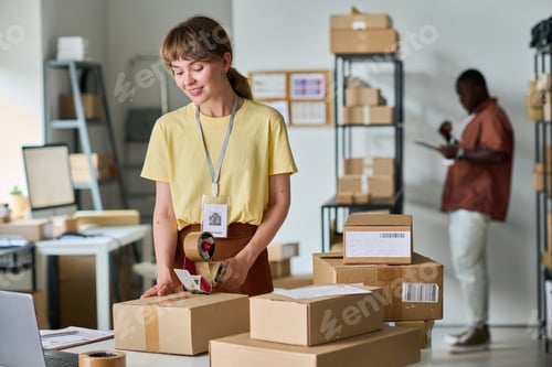 Preview: Young smiling woman in yellow t-shirt sealing cardboard box with cellotape