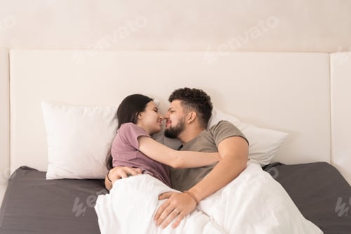 Preview: Happy young affectionate couple in t-shirts lying in embrace under white blanket on large double bed