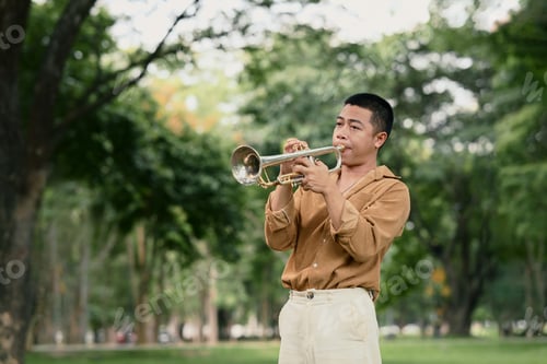Preview: Young man playing trumpet in a park surrounded by lush green trees on a bright day