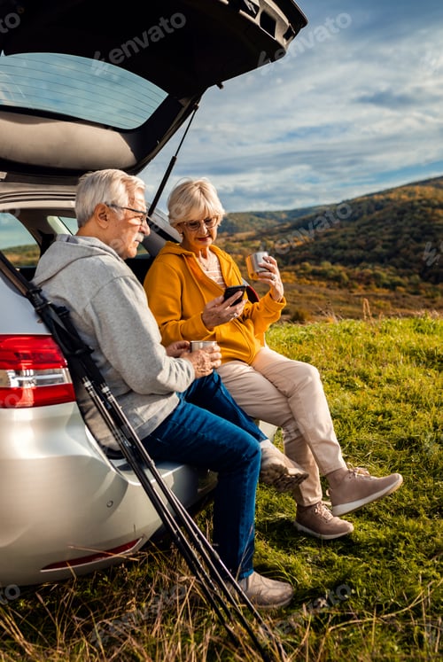 Preview: Senior couple sitting against the car, resting after hiking in countryside.