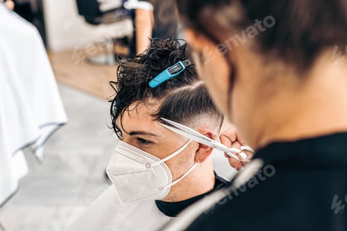 Preview: Young man with mask sitting while a woman cut his hair in a hairdresser