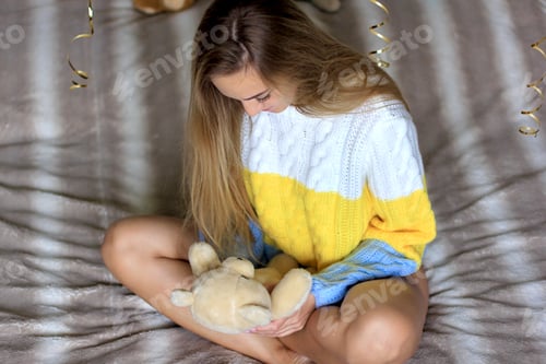 Preview: Young beautiful woman in colorblock cozy sweater resting on the bed at home. Teddy bear in hands.