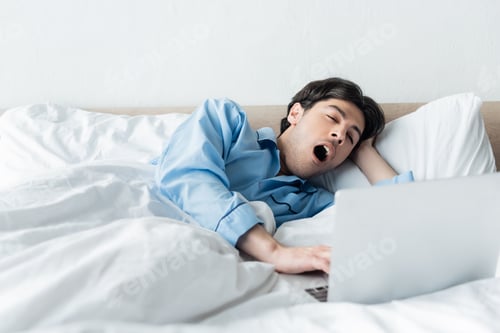 Preview: sleepy man yawning near laptop while lying in bed in morning