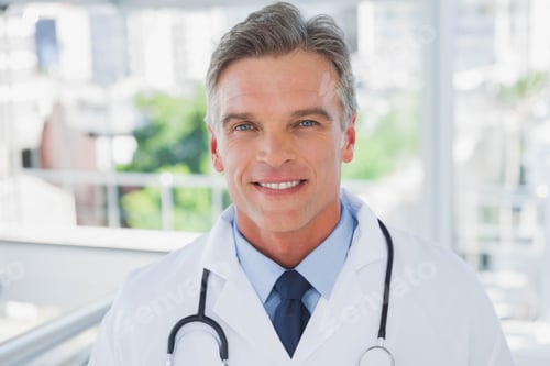 Preview: Male physician wearing white coat and smiling while standing in clinic corridor holding stethoscope