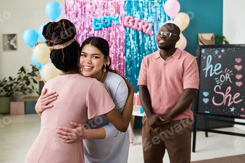 Preview: Young adult Asian woman hugging her pregnant friend at gender reveal party