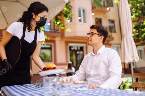 Preview: Waitress in protective mask serving delicious food for man in cozy restaurant.