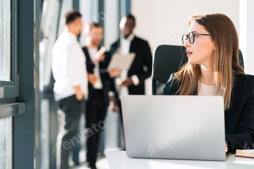 Vista previa: La secretaria pensativa con gafas se sienta frente a la computadora portátil y mira hacia otro lado