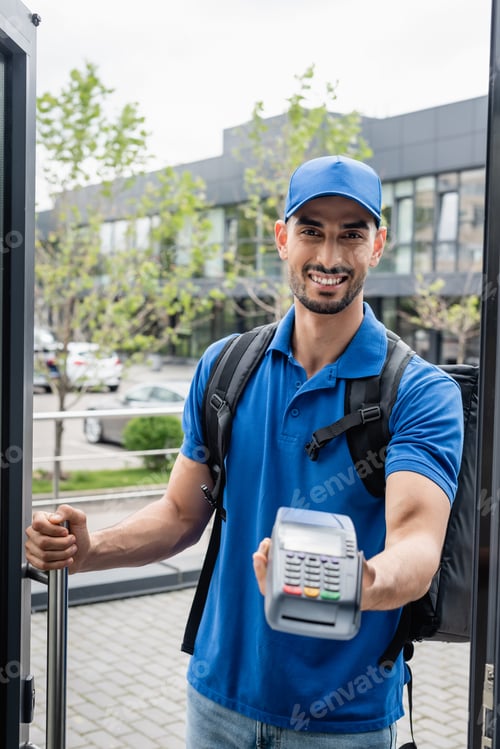 Preview: Cheerful muslim courier holding blurred payment terminal near door