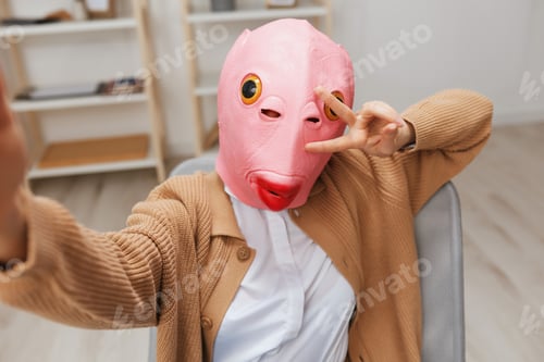 Preview: Funny blonde lady in pink fish mask warm sweater doing selfie use show v sign gesture phone in
