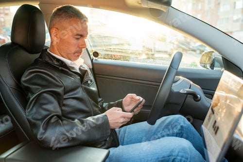 Preview: Contemporary man texting in smartphone while sitting in electric car