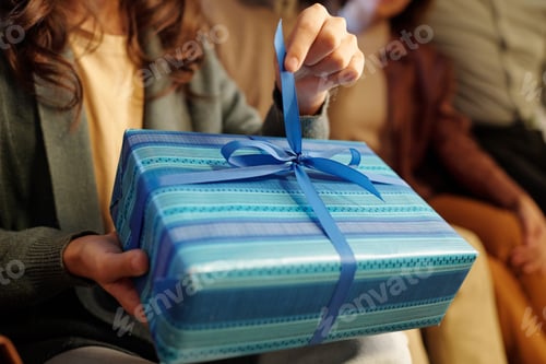Preview: Hand of youthful girl pulling thin blue silk ribbon on top of giftbox