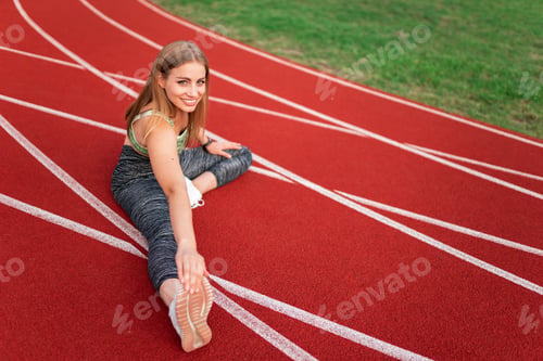 Preview: Positive sports girl doing stretching while sitting on the stadium track