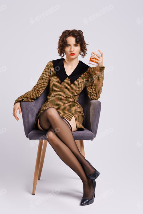 Preview: Woman with curly hair in dress and stilettos is sitting in grey studio with a glass of whiskey