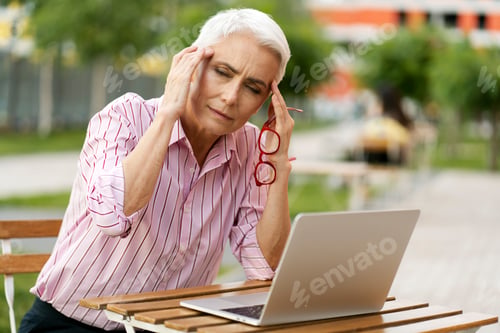 Preview: Tired senior woman holding red glasses, having headache outdoors. Health problems, migraines