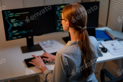 Preview: Rear view of young female software engineer working at night in office.
