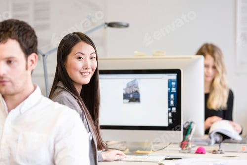 Preview: Mid adult woman sitting at computer looking over shoulder at camera smiling