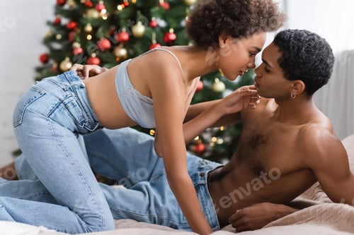 Preview: side view of passionate african american woman in bra and jeans near man in bedroom with christmas