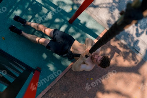 Preview: sportsman with a bare torso pulls up on a rope during outdoor training on the sports ground