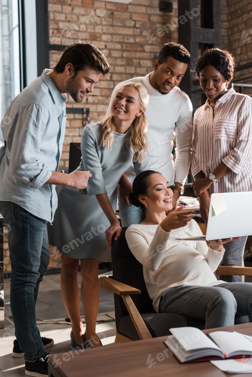 Preview: cheerful multicultural businesspeople standing near happy asian colleague using laptop
