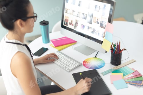 Preview: Rear view of attentive Asian female graphic designer using graphic tablet at desk in a modern office