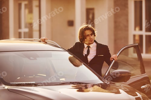 Preview: Portrait of handsome young businessman in black suit and tie outdoors near modern car in the city