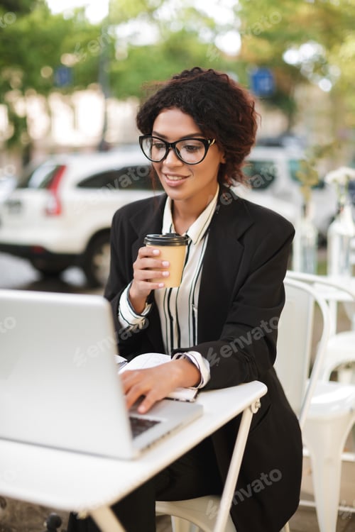 Preview: Smiling African American girl in glasses sitting at the table of cafe and working on her laptop