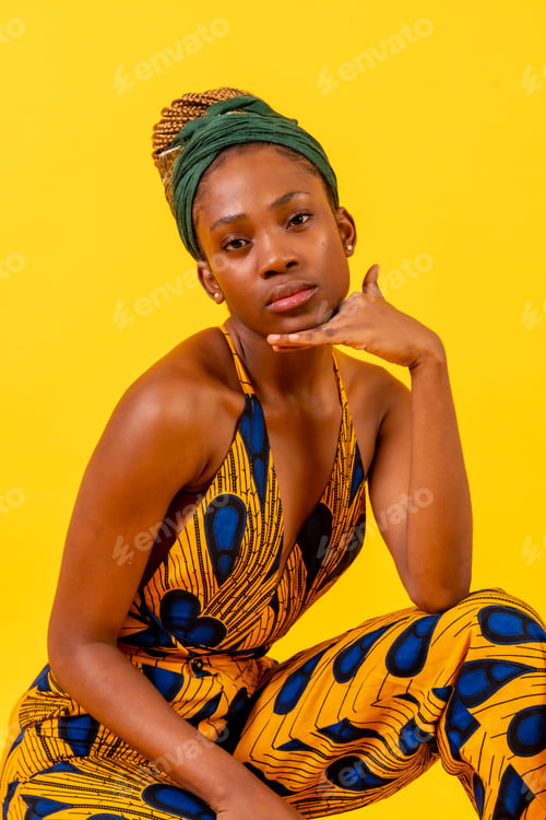 Preview: African young woman in traditional dress on yellow background sitting in studio looking at camera