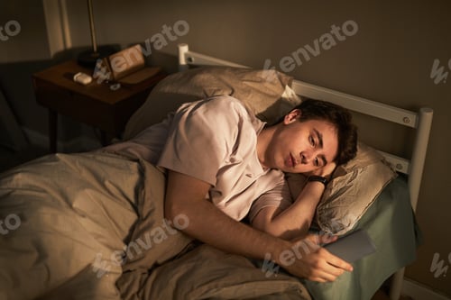Preview: Young Caucasian Man Lying in Bed Feeling Sad While Holding Smartphone