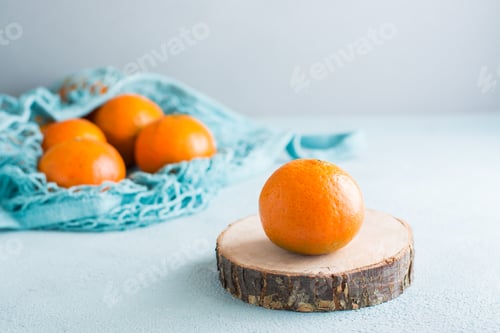 Preview: Ripe tangerine on a cut of a tree and tangerines in a mesh bag on a blue background.