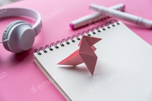 Preview: Selective focus shot of swan origami with notebook, markers, and headphones on pink background